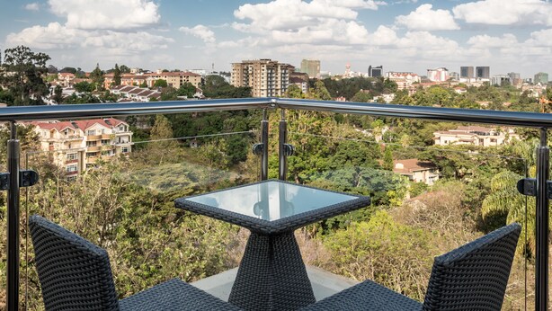 Un balcon avec une table en verre et des chaises en osier, donnant sur un paysage urbain avec des immeubles et des arbres.