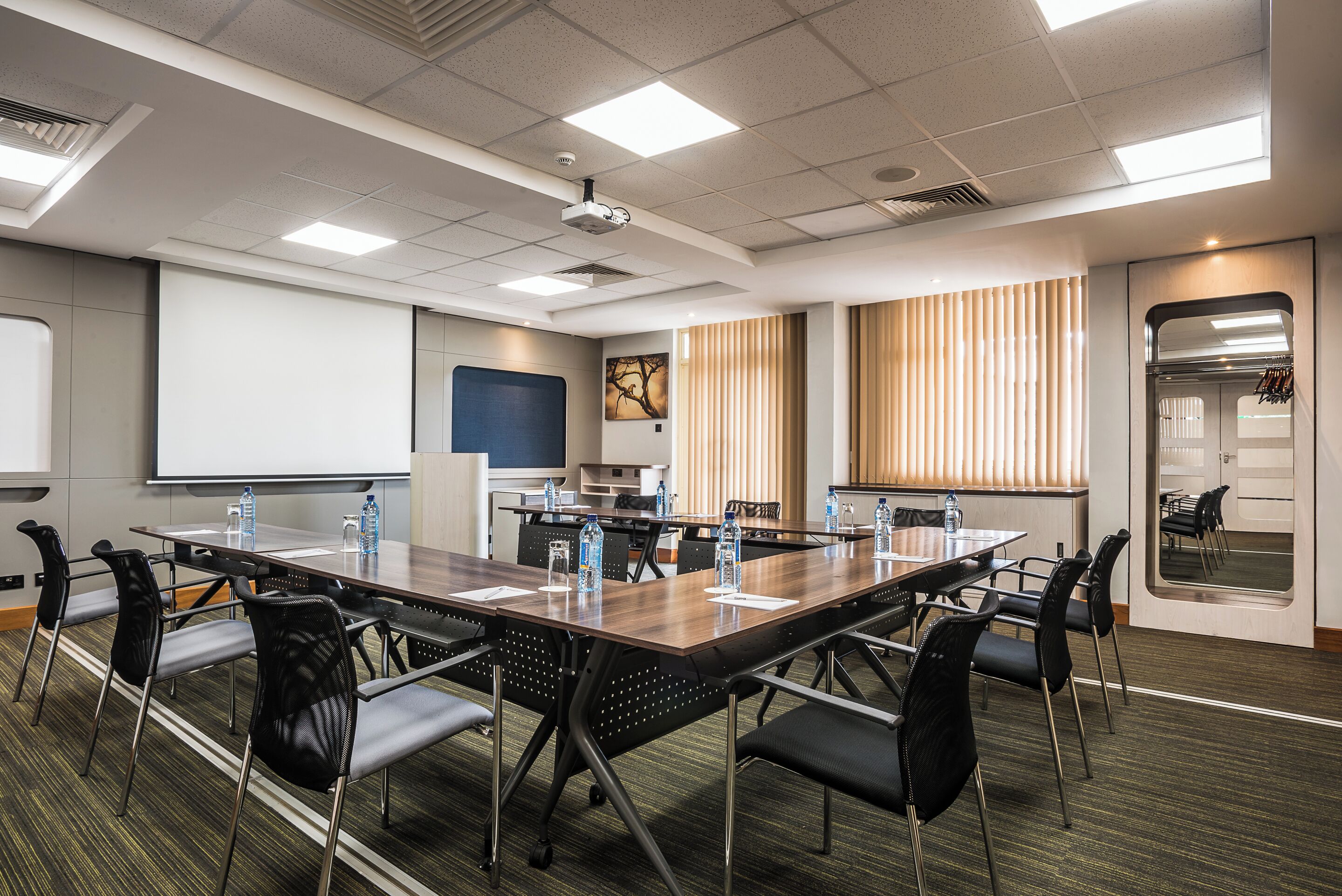 Sala de conferências com uma mesa comprida, cadeiras, garrafas de água, tela de projetor e uma grande janela com persianas.