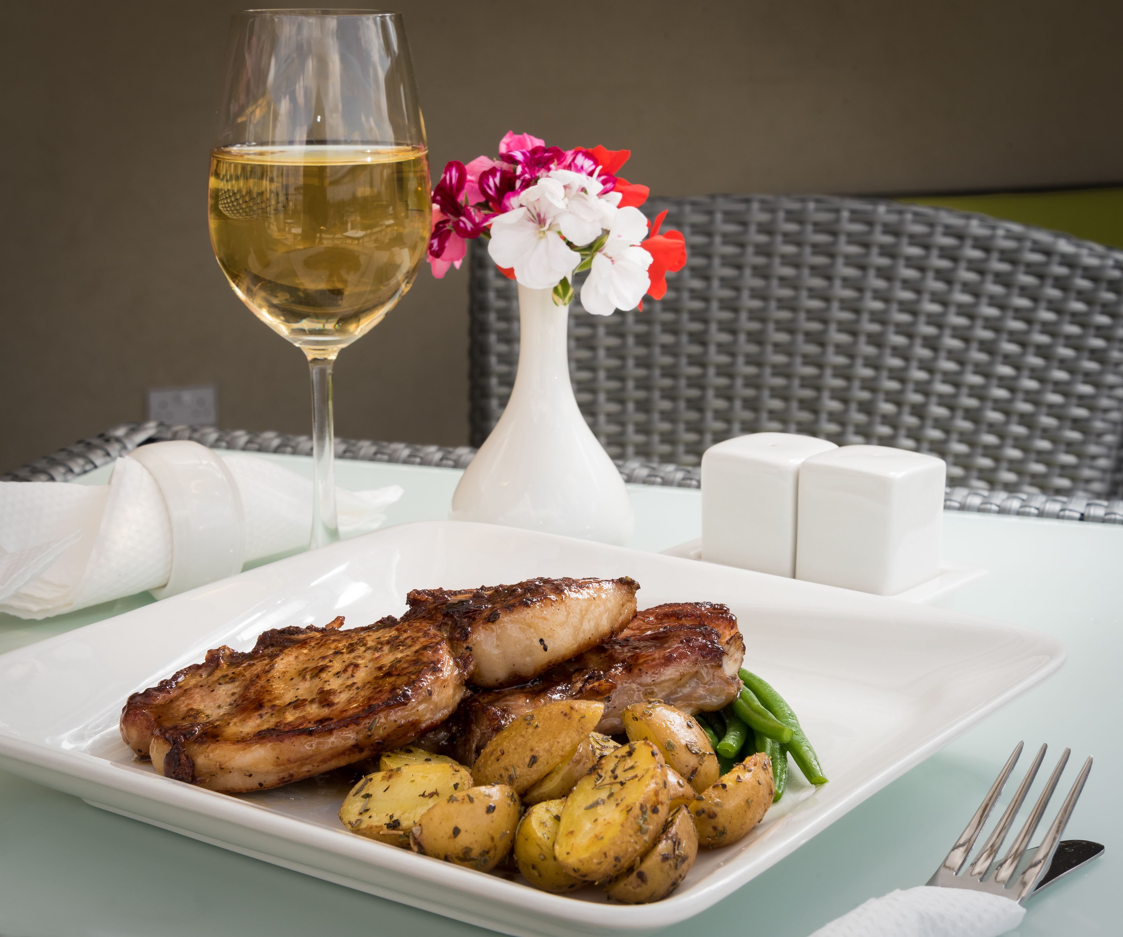 A plate of grilled meat, roasted potatoes, and green beans on a white table with a glass of white wine and a vase of flowers.