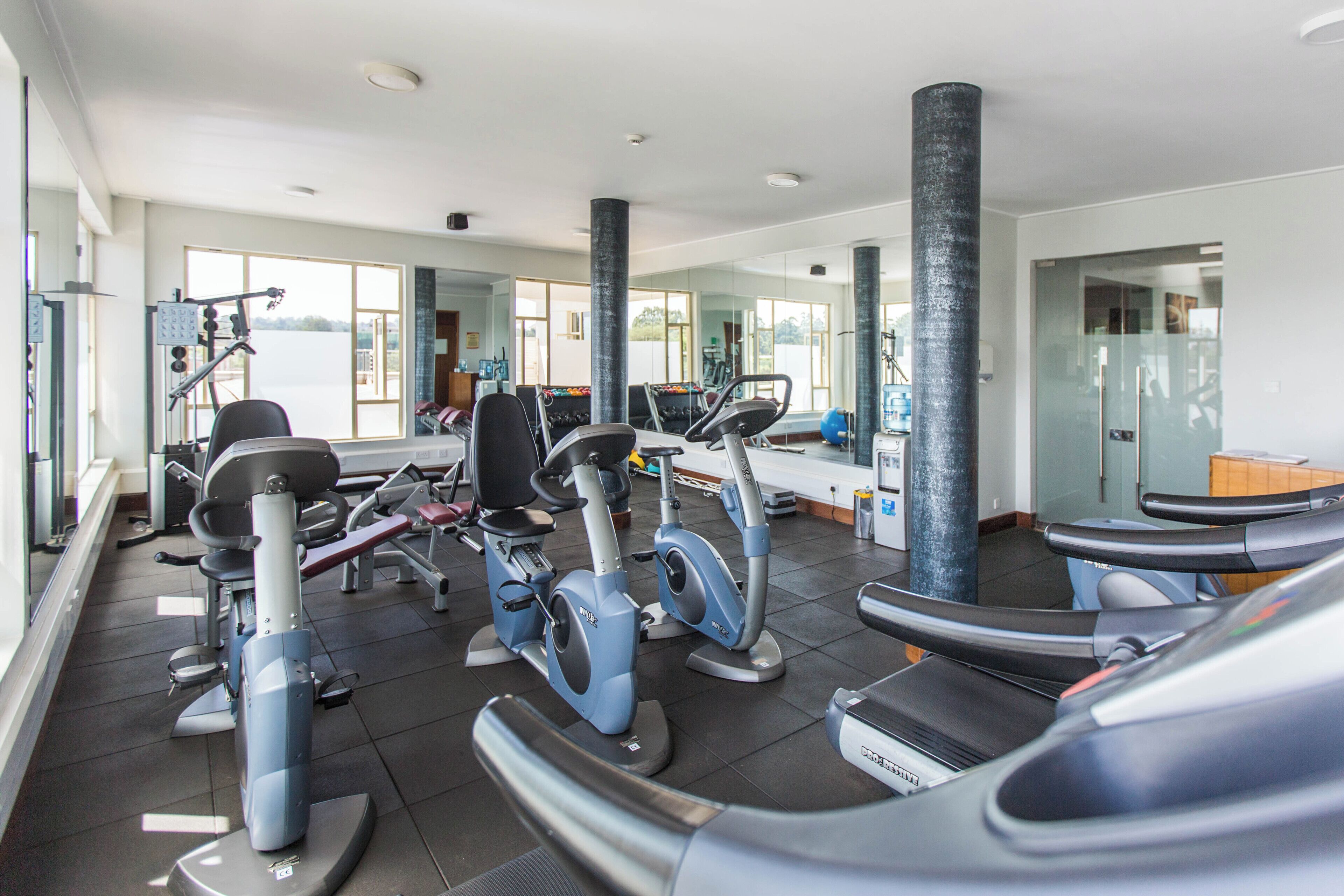 Un gimnasio con varios equipos de ejercicio, incluyendo cintas de correr y máquinas de pesas, y ventanas amplias que permiten la entrada de luz natural.