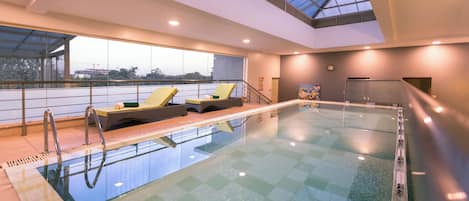 An indoor swimming pool with a glass floor, lounge chairs, and a view of the outside.