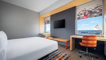 Quarto de hotel moderno com uma cama grande, uma mesa, uma cadeira, uma TV e uma janela com vista para a cidade.