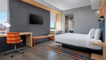 A modern hotel room with a large bed, a desk with a chair, a flat-screen TV, and a city view.