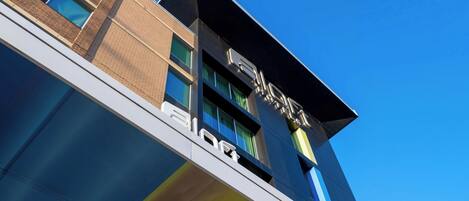 Un edificio moderno con un llamativo letrero 'AIO' en la fachada, ventanas amplias y un cielo azul despejado al fondo.