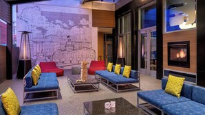 A modern lounge area with blue sofas, yellow and red cushions, a glass coffee table, and a large mural of a cityscape. - Aloft by Marriott Columbia Downtown (Columbia)