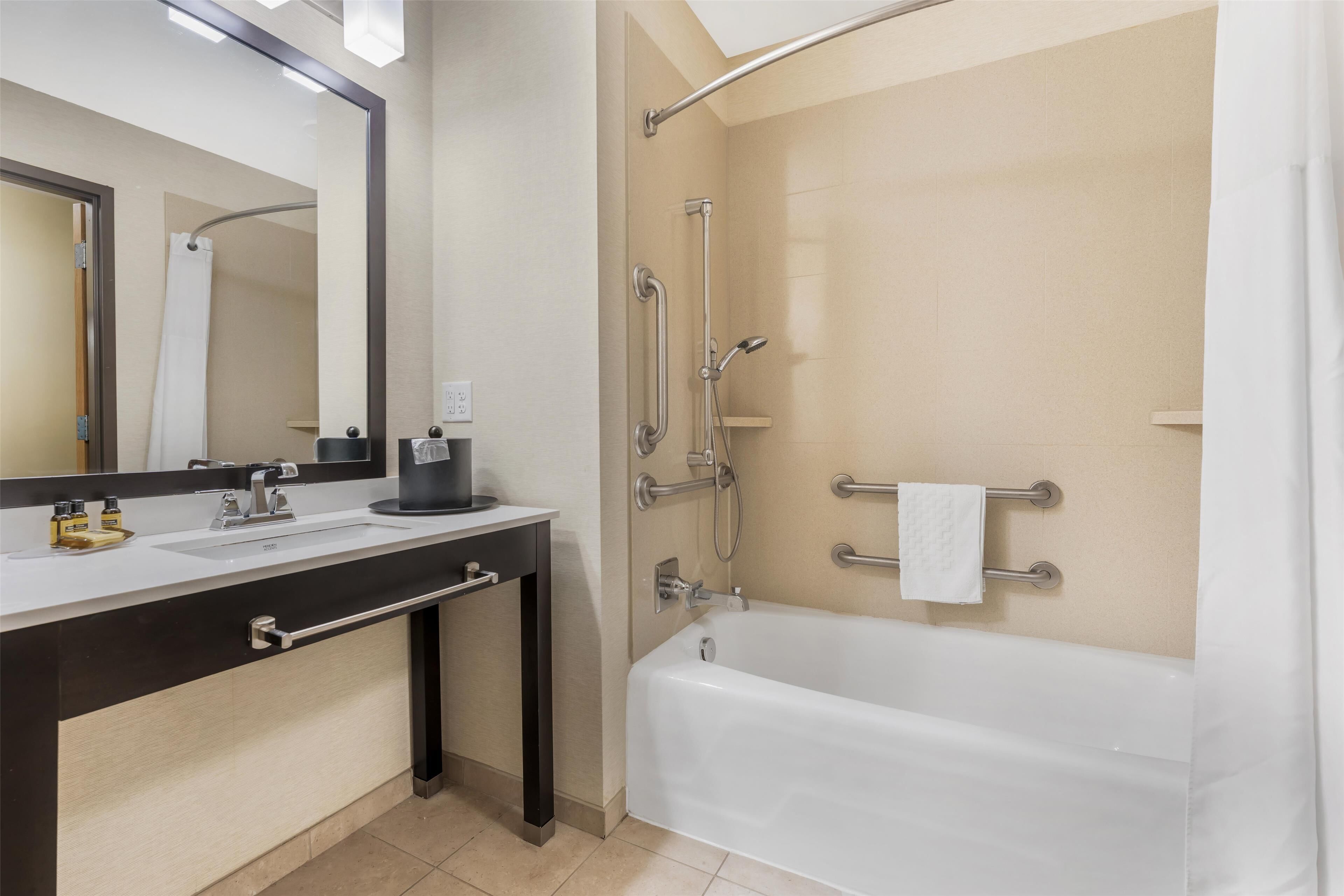 Une salle de bain moderne équipée d’une baignoire, d’une douche avec des barres d’appui, d’un meuble-lavabo avec un miroir et d’un porte-serviettes.
