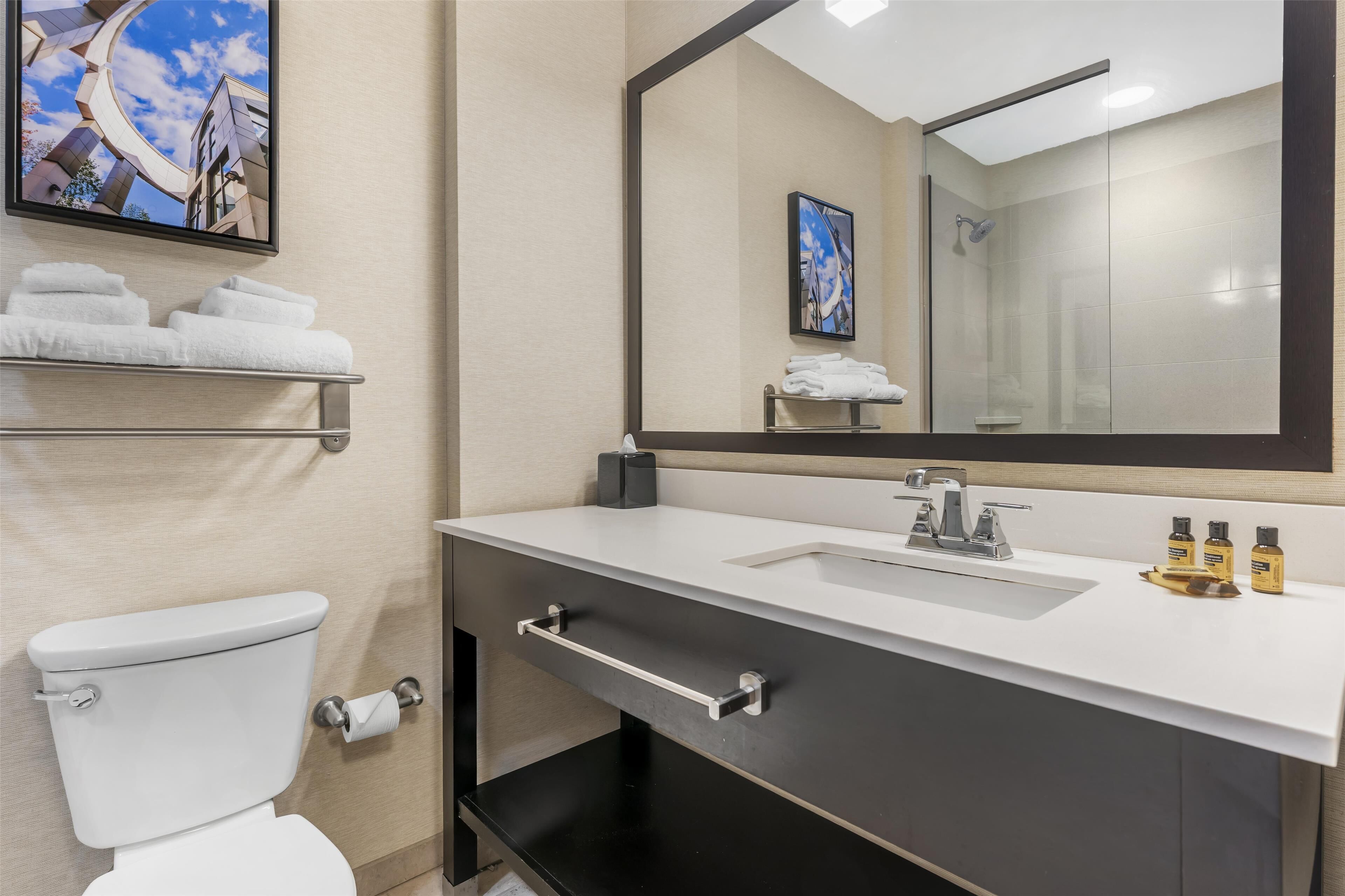 A modern bathroom with a toilet, sink, and mirror. There are towels on a shelf, and toiletries on the sink.