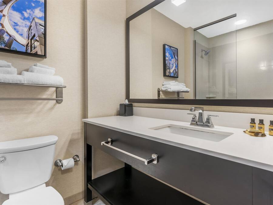 A modern bathroom with a toilet, sink, and mirror. There are towels on a shelf, and toiletries on the sink.