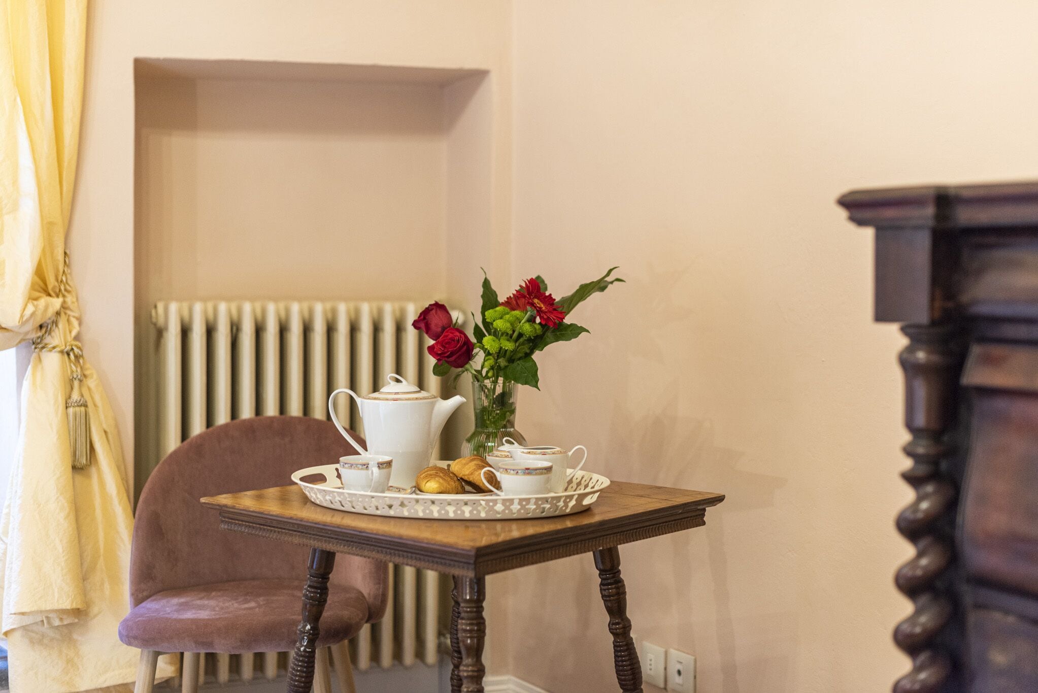 A dining table with a teapot, cups, and a tray with pastries, set against a wall with a radiator and a chair.