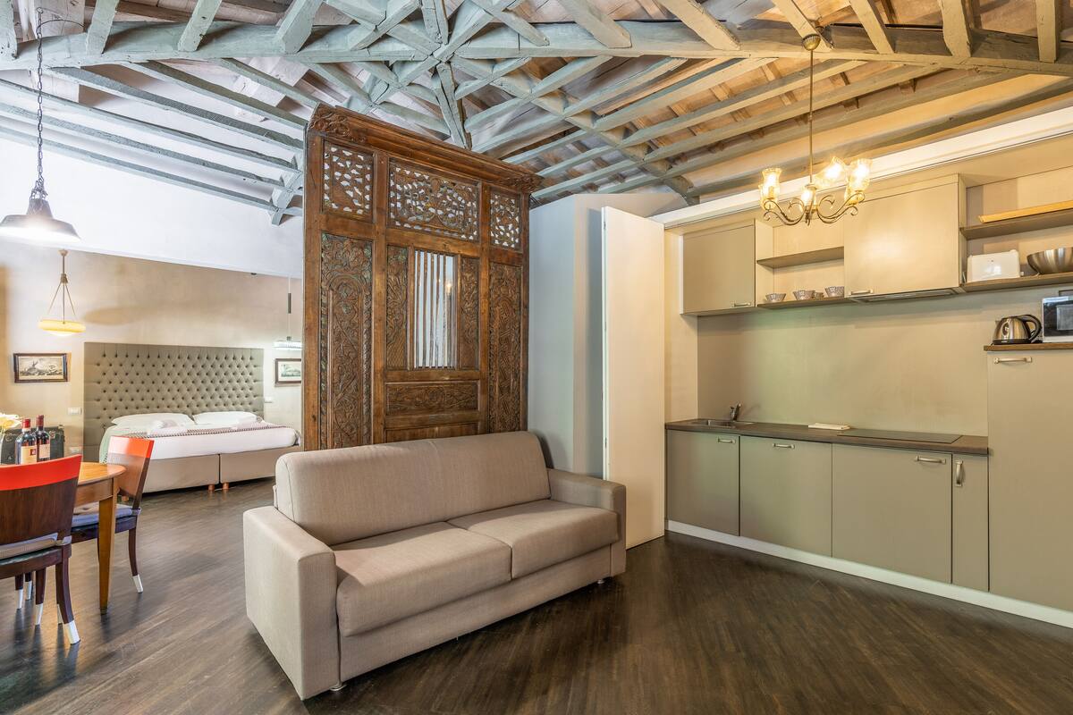 A modern living room with a sofa, dining table, and chairs, a kitchen area with cabinets and appliances, and a decorative wooden partition.