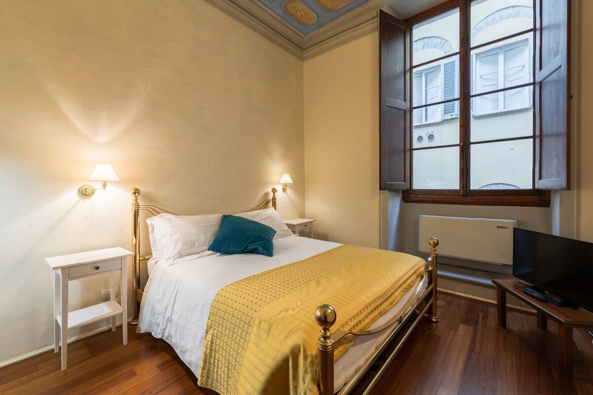 A bedroom with a bed, bedside tables, a television, and a window with a view of buildings.