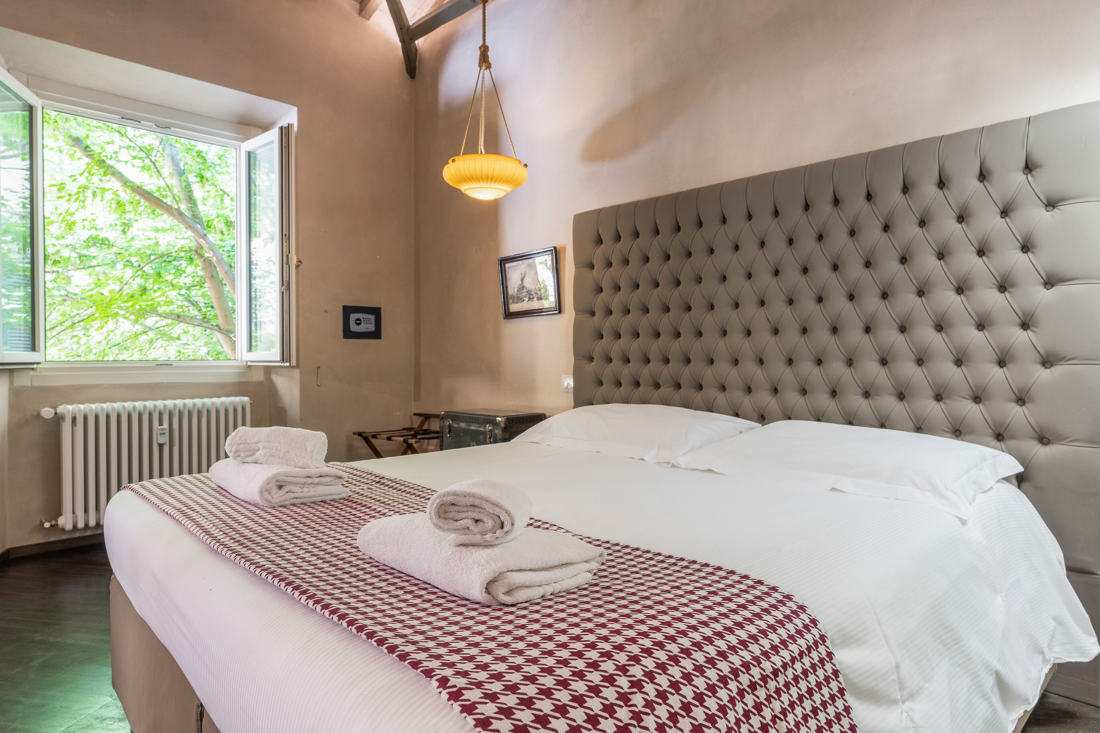 A neatly made bed with a patterned bedspread, a tufted headboard, and a window with a view of trees.