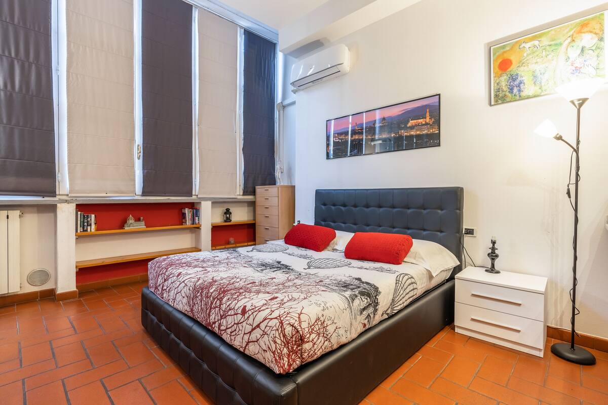 A bedroom with a bed, a nightstand, a lamp, a shelf with books, and a window with curtains.
