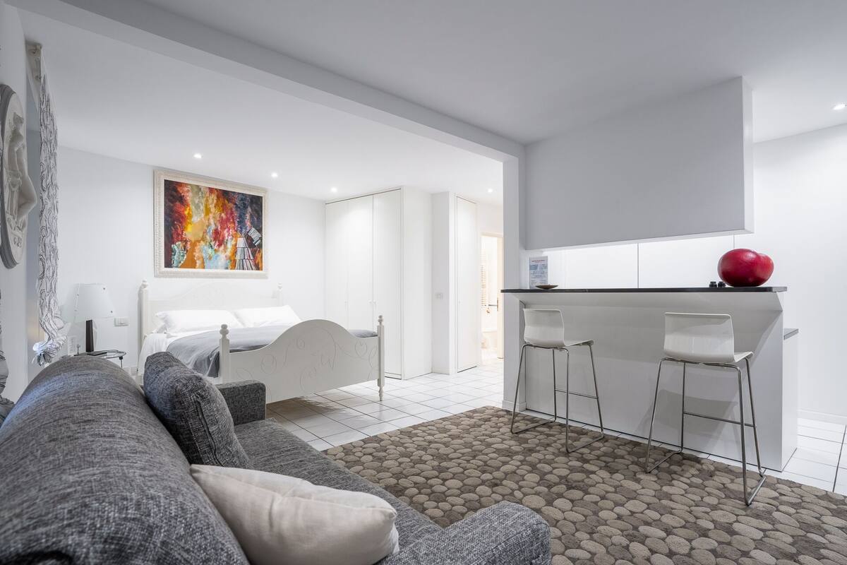A modern, minimalist bedroom with a bed, a gray sofa, and a colorful abstract painting.