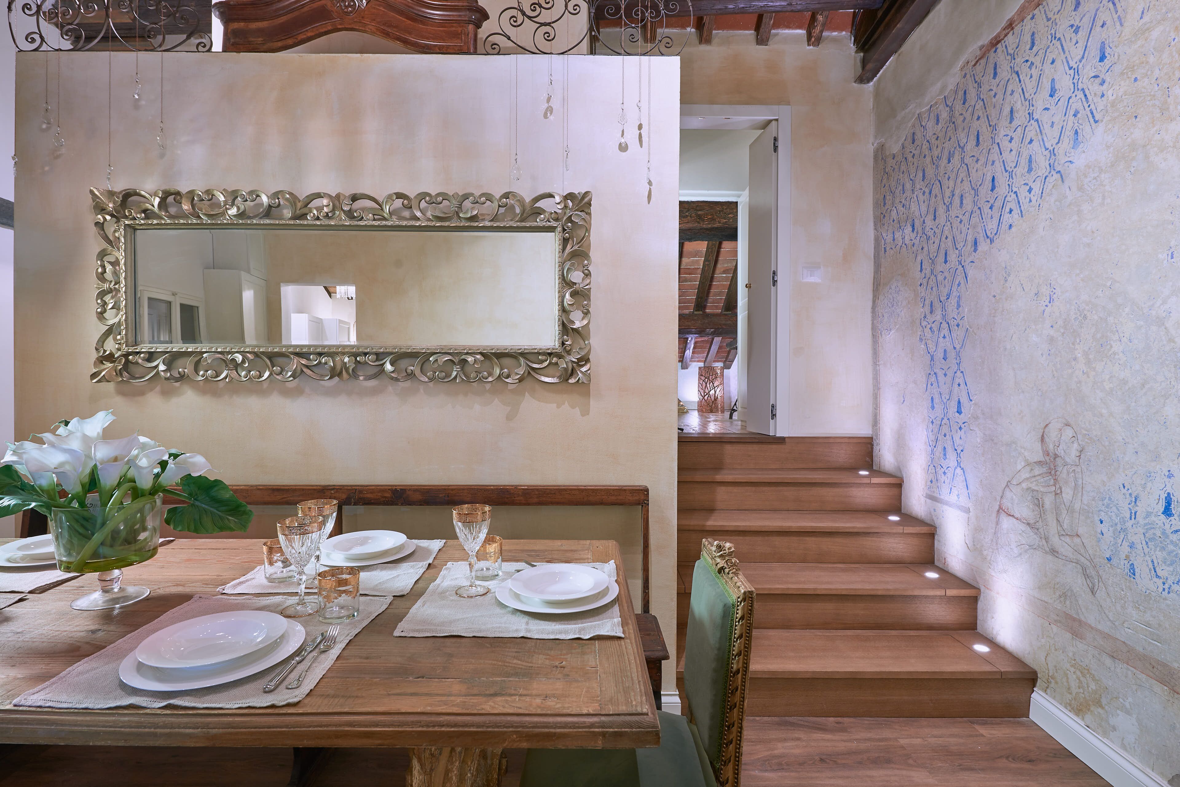 A dining area with a wooden table set for four, a mirror above it, and a staircase with a patterned wall to the right.