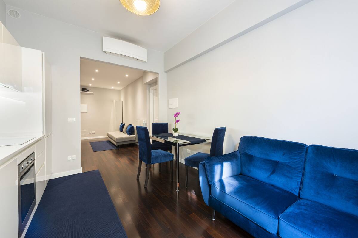 A modern living room with a blue sofa, a dining table with chairs, and a kitchen area with a stove.