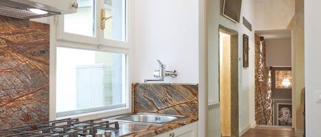 Una cocina con estufa a gas, pileta de baño y encimera de mármol. Hay alacenas y una ventana sobre la pileta.