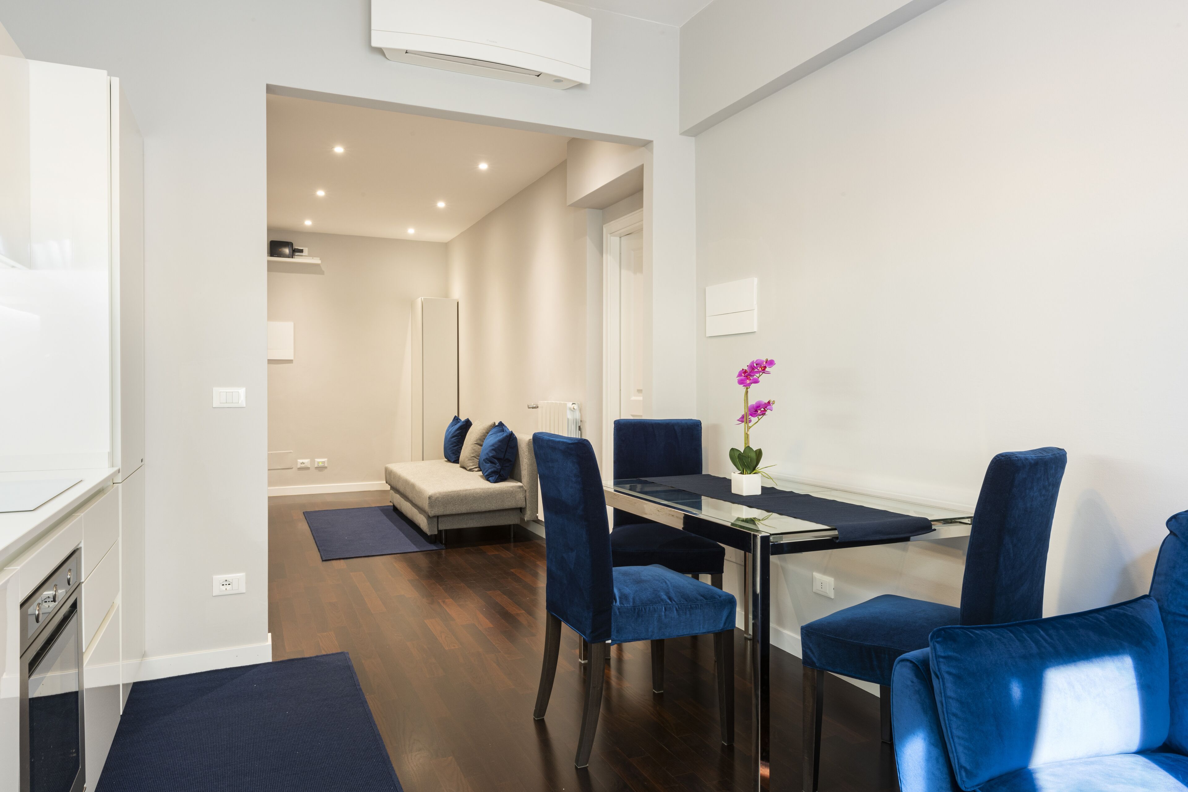 Uma sala de jantar moderna com mesa de vidro, cadeiras estofadas em azul e uma orquídea em vaso.