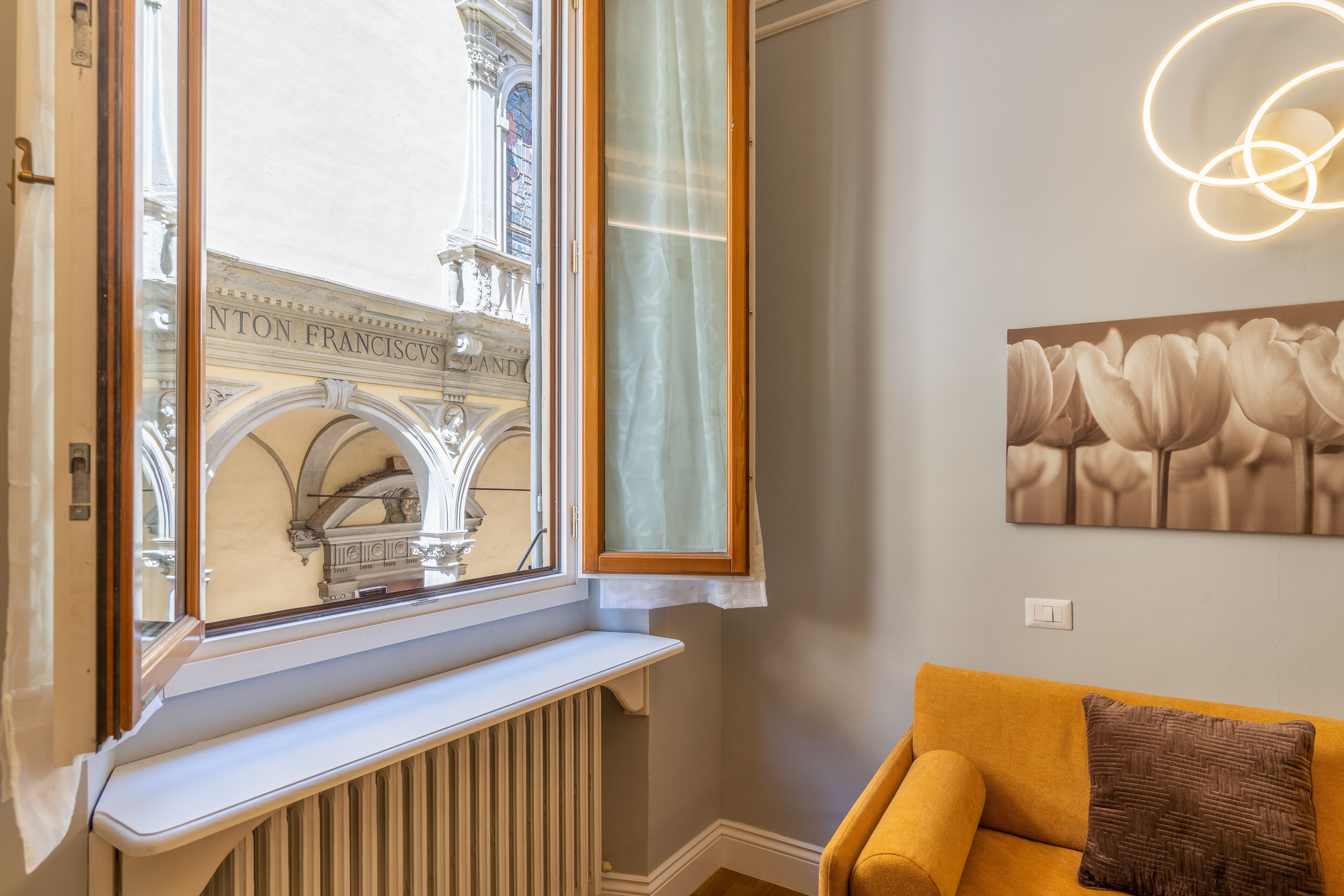 A room with a window overlooking a historic building, a radiator below the window, a yellow sofa with a patterned pillow, and a wall-mounted light fixture.