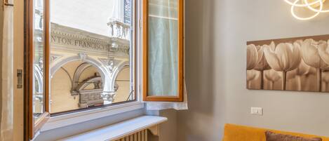 A room with a window overlooking a historic building, a radiator below the window, a yellow sofa with a patterned pillow, and a wall-mounted light fixture.