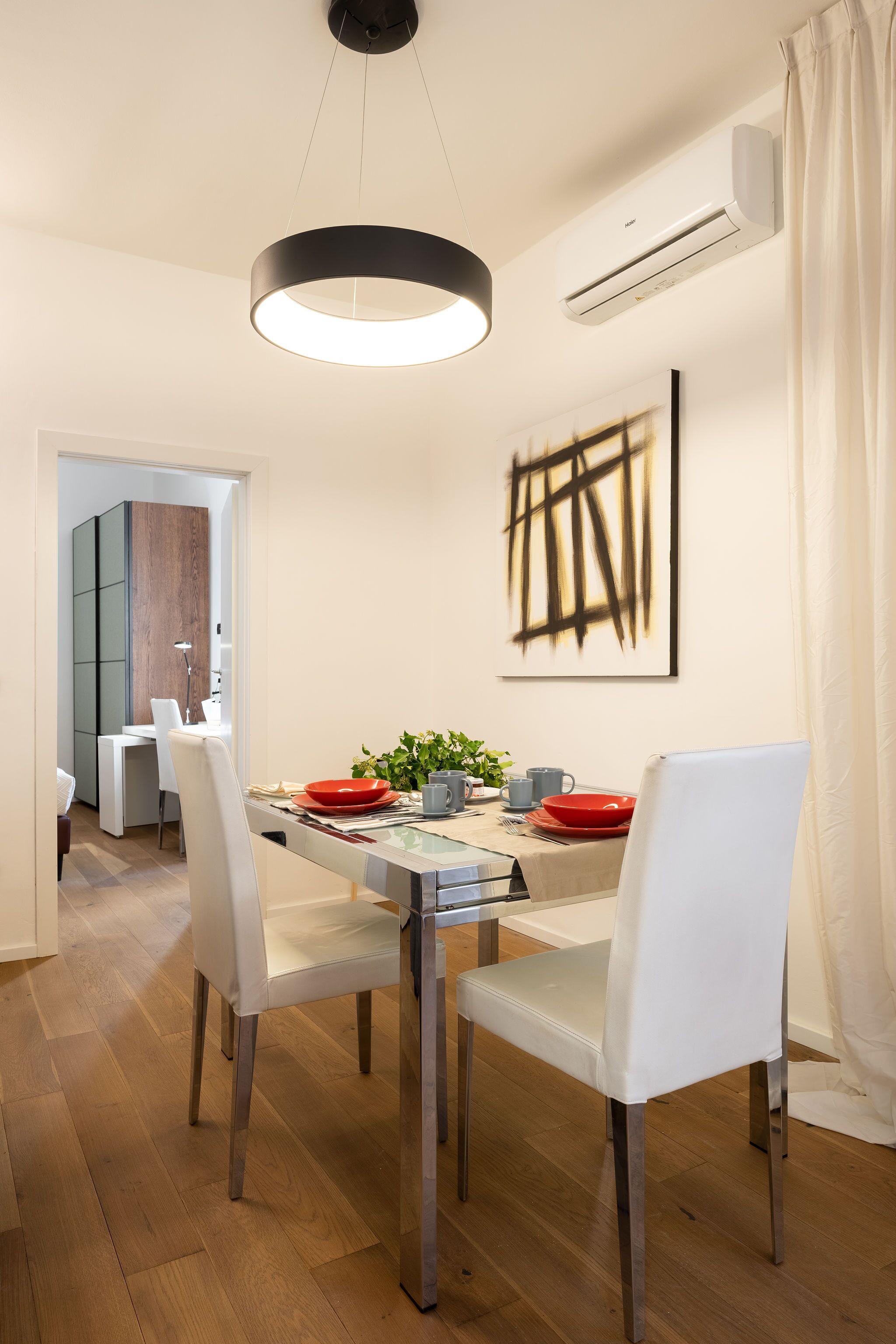 Un comedor moderno con una mesa blanca, cuatro sillas blancas, un cuenco rojo y una lámpara colgante negra.