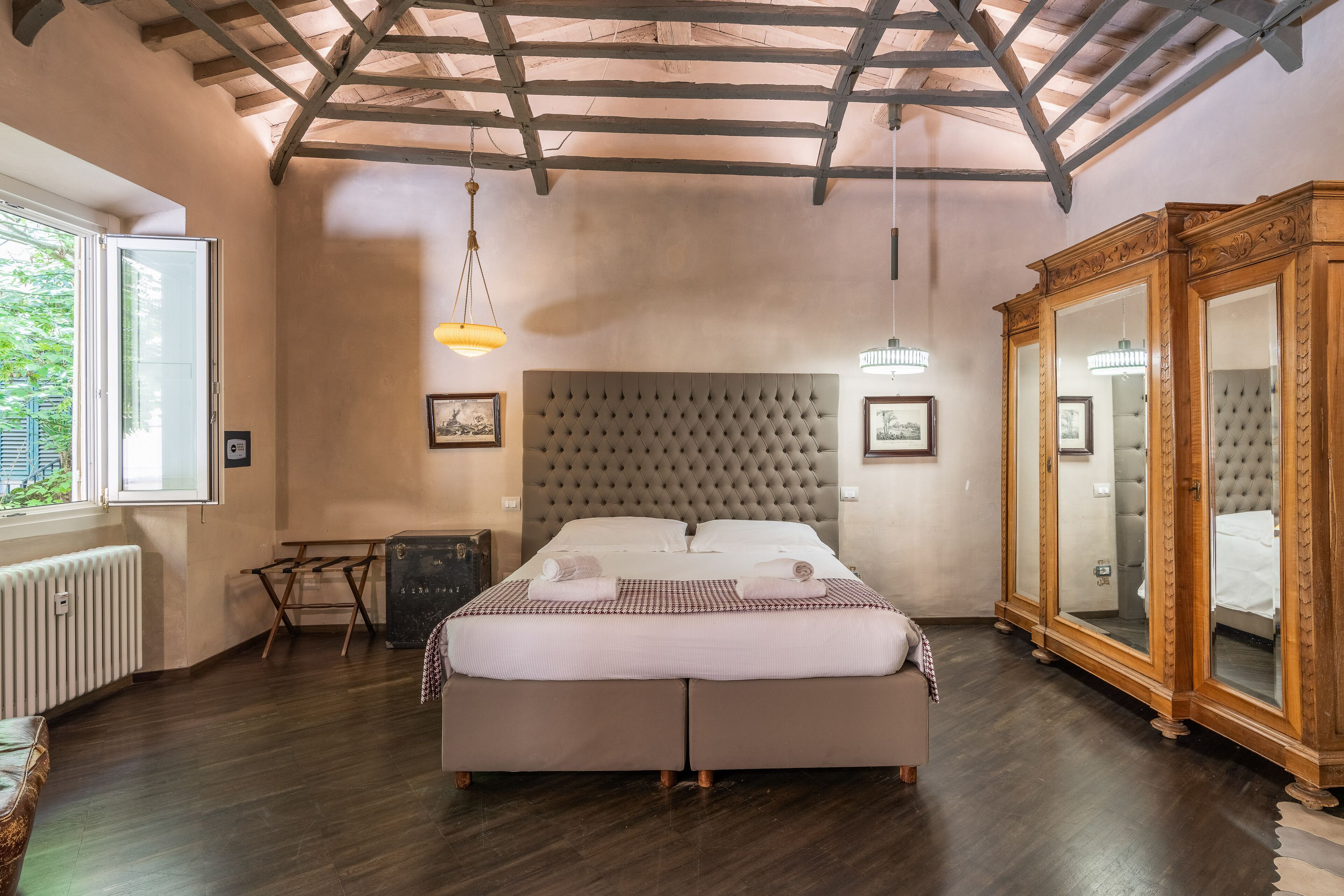 Un dormitorio con una cama grande, vigas de madera en el techo, un radiador, un armario y una ventana con vistas al verdor.