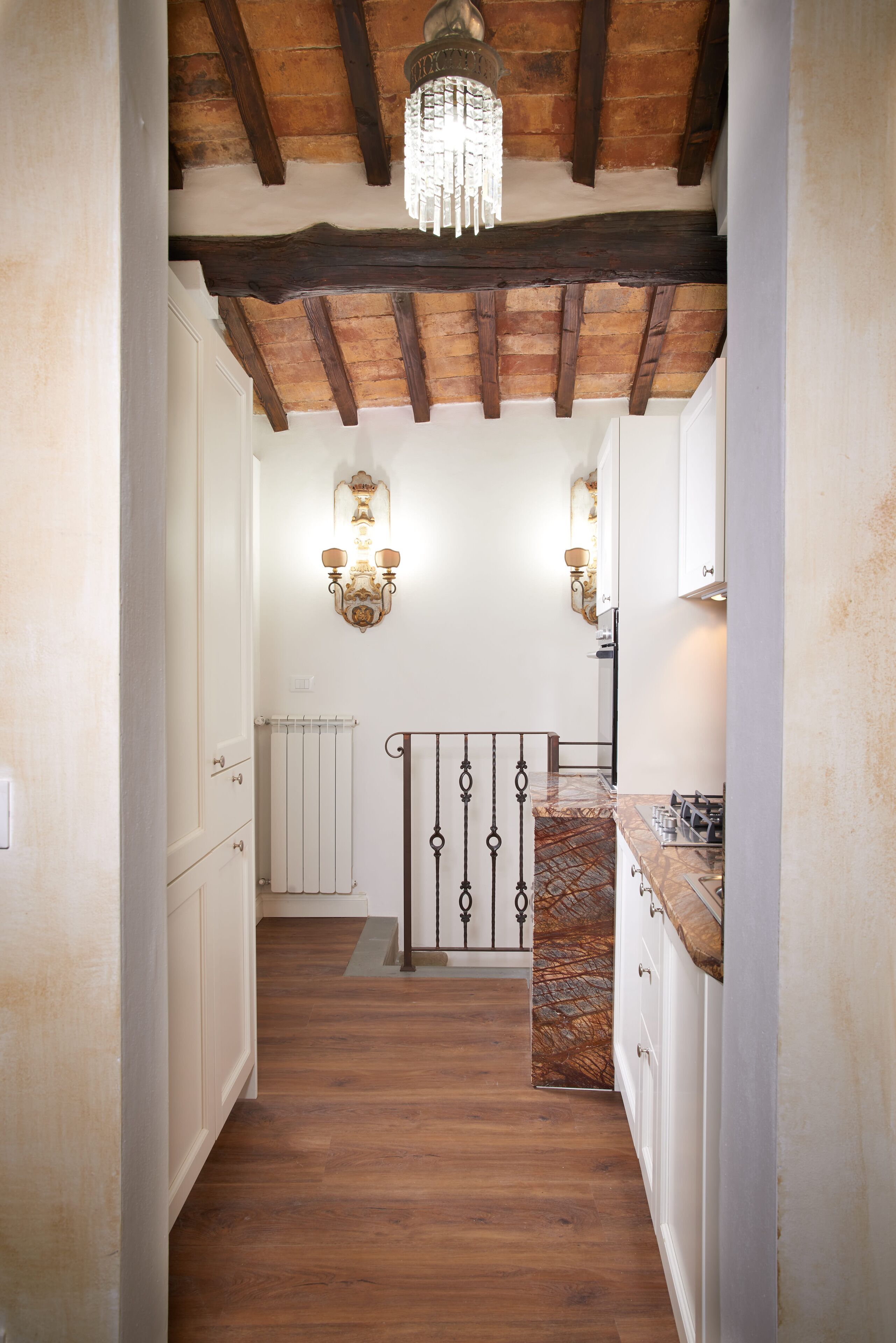 Un pasillo estrecho con suelo de madera, paredes blancas y techo con vigas a la vista. A la izquierda hay un radiador y a la derecha, una zona de cocina con encimera de mármol y una estufa.
