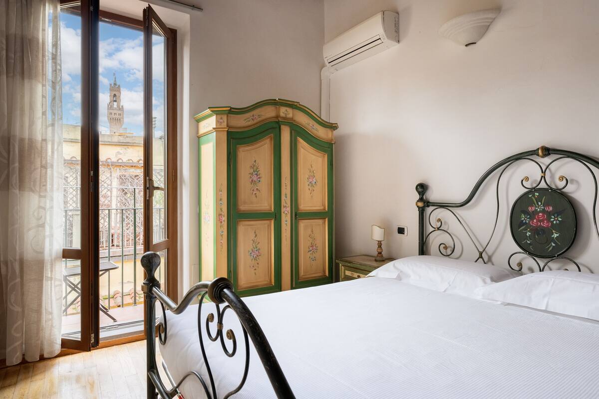 A bedroom with a bed, a wardrobe, a nightstand, and a view of a tower through a window.