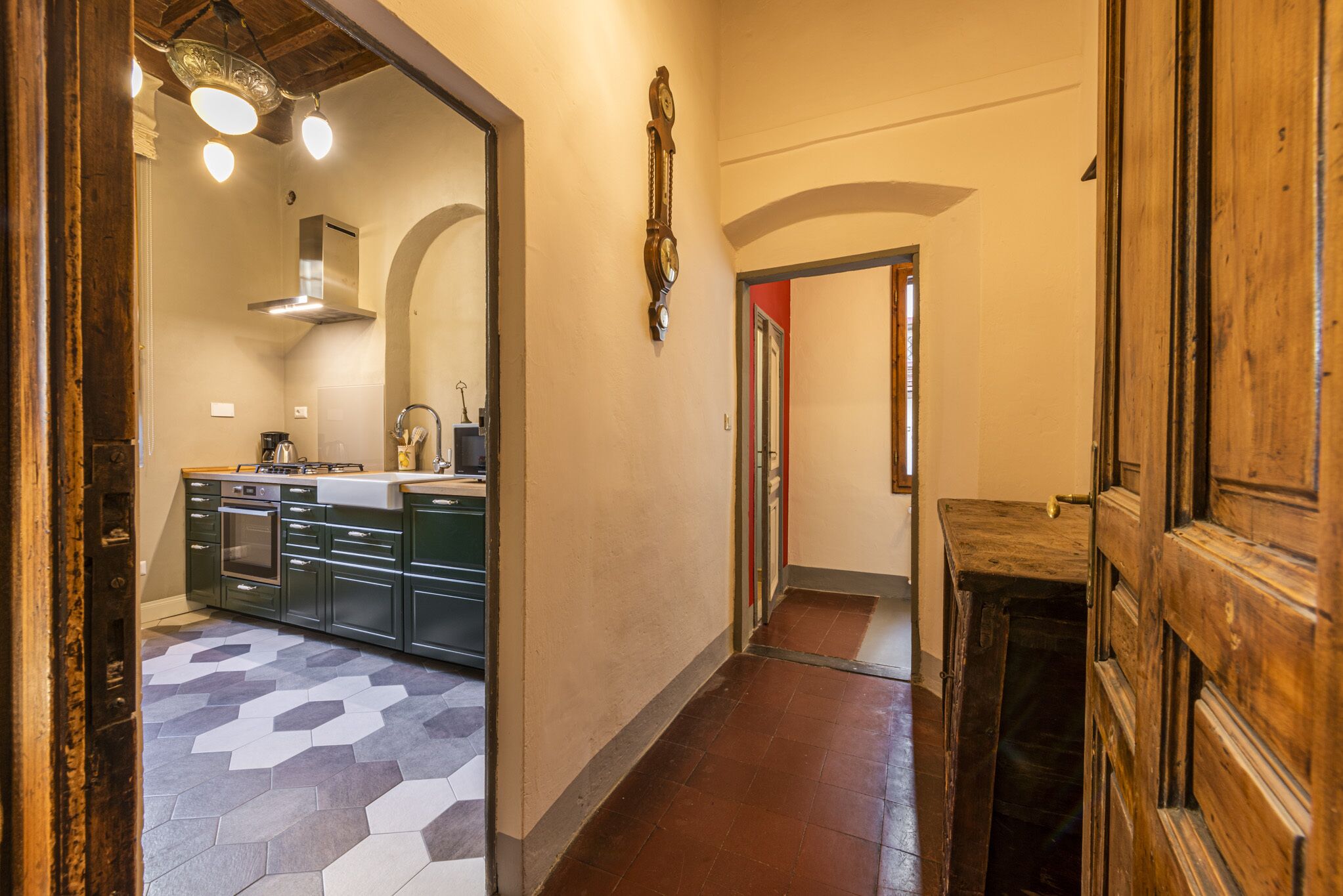 A hallway with a kitchen area featuring cabinets, a stove, and a sink. There is a wooden door on the right and a mirror on the left.