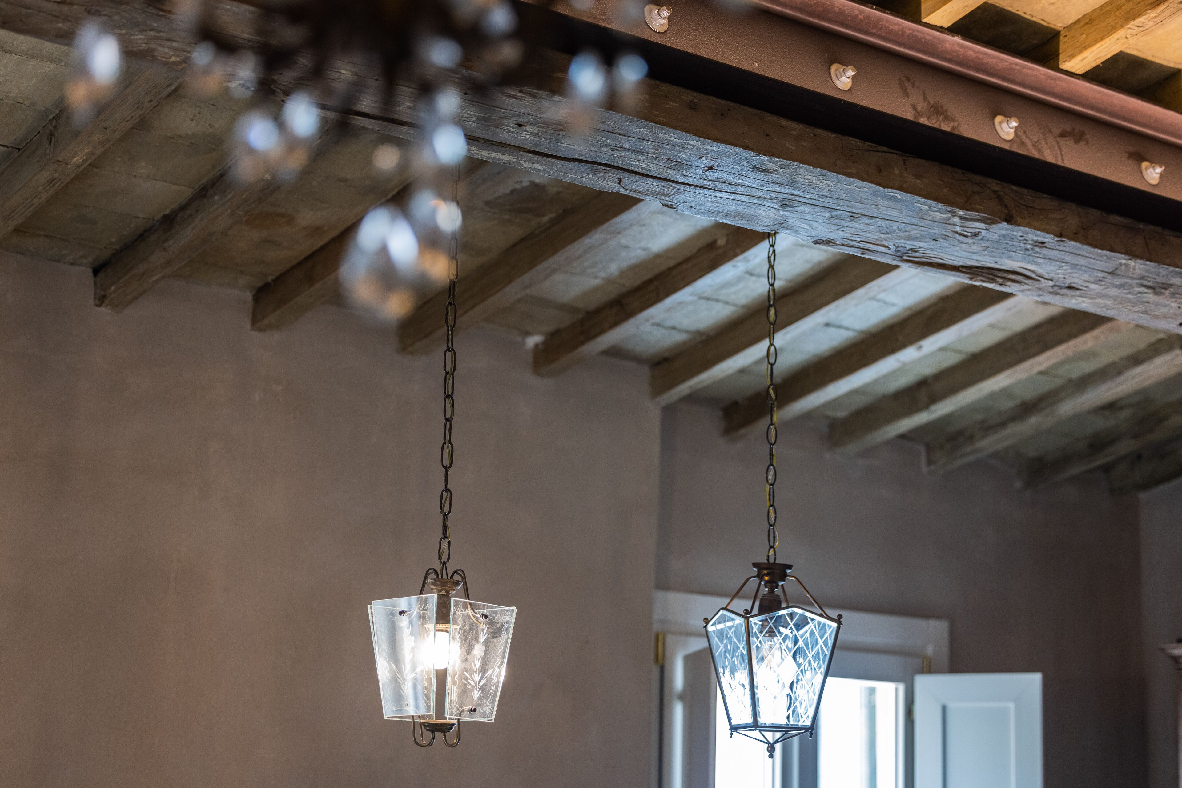 Dos lámparas colgantes de cristal con diseño tipo farol en una habitación con vigas de madera a la vista y una puerta blanca al fondo.