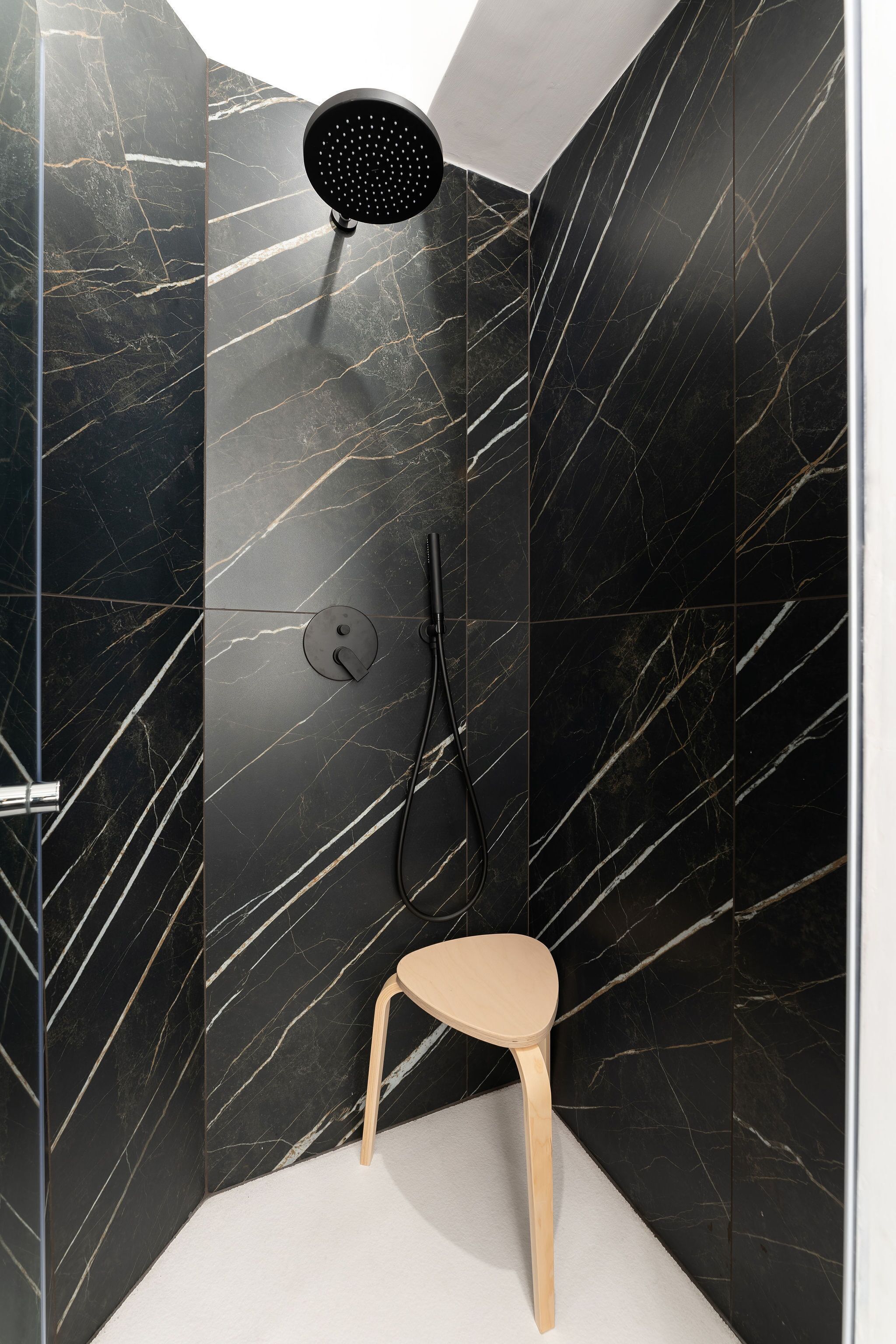 A modern shower with a handheld showerhead and a handheld shower attachment, a single wooden chair, and a marble wall with white veining.