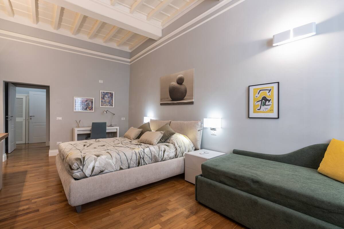 A modern bedroom with a bed, a green sofa, a desk with a chair, and artwork on the walls.