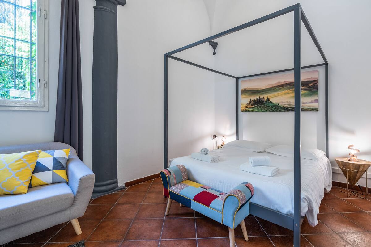 A four-poster bed with a view of a landscape painting, a sofa with patterned cushions, and a small wooden side table with a lamp.