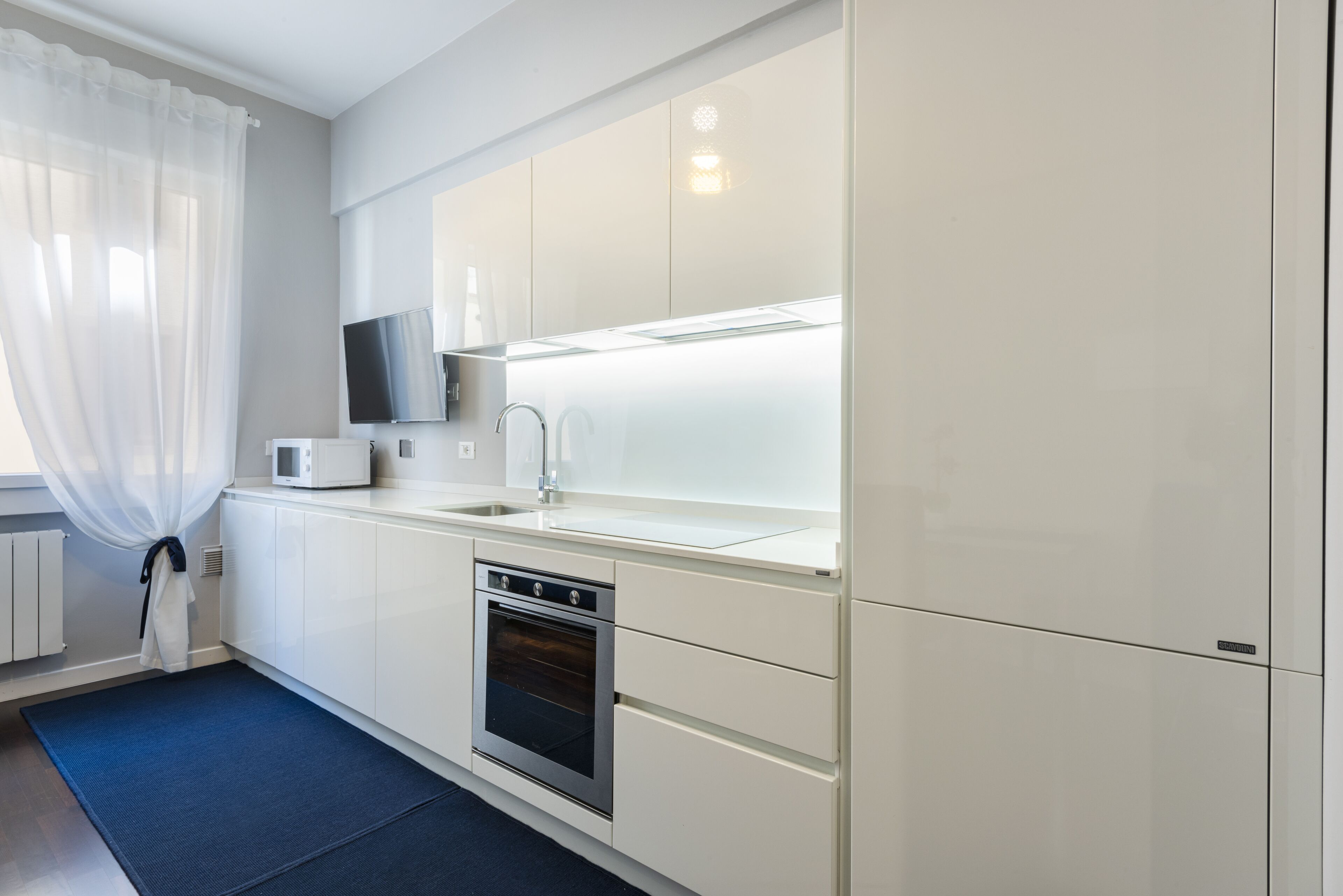 A modern kitchen with white cabinets, a stainless steel oven, a microwave, and a flat-screen TV mounted above the sink.
