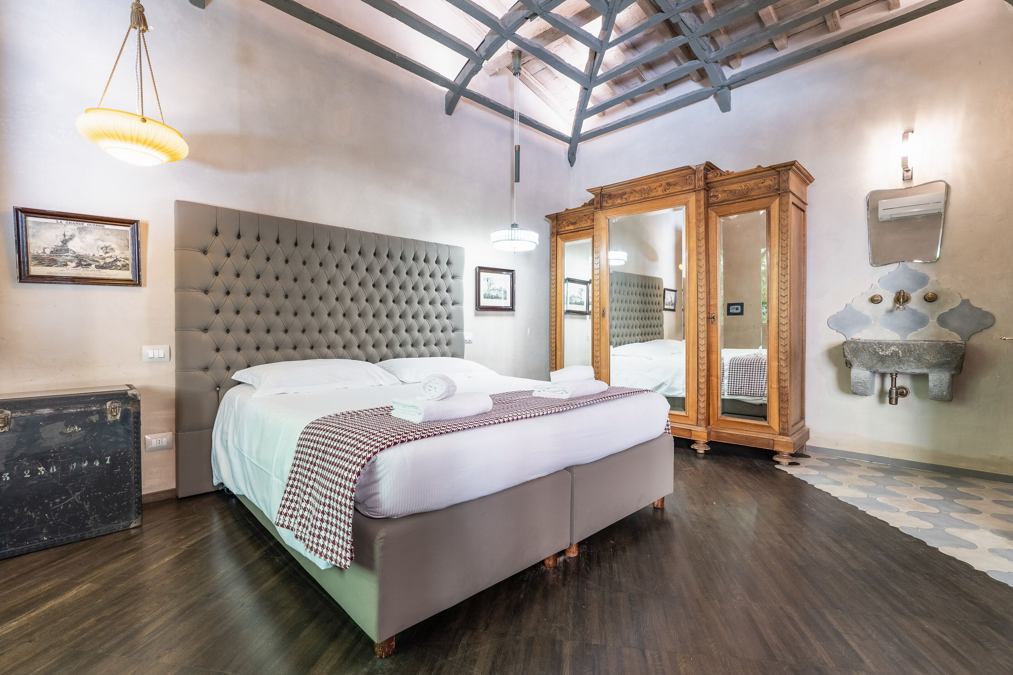 Un dormitorio con cabecera capitonada, cama con ropa de cama blanca, armario de madera, espejo y lavamanos de piedra.