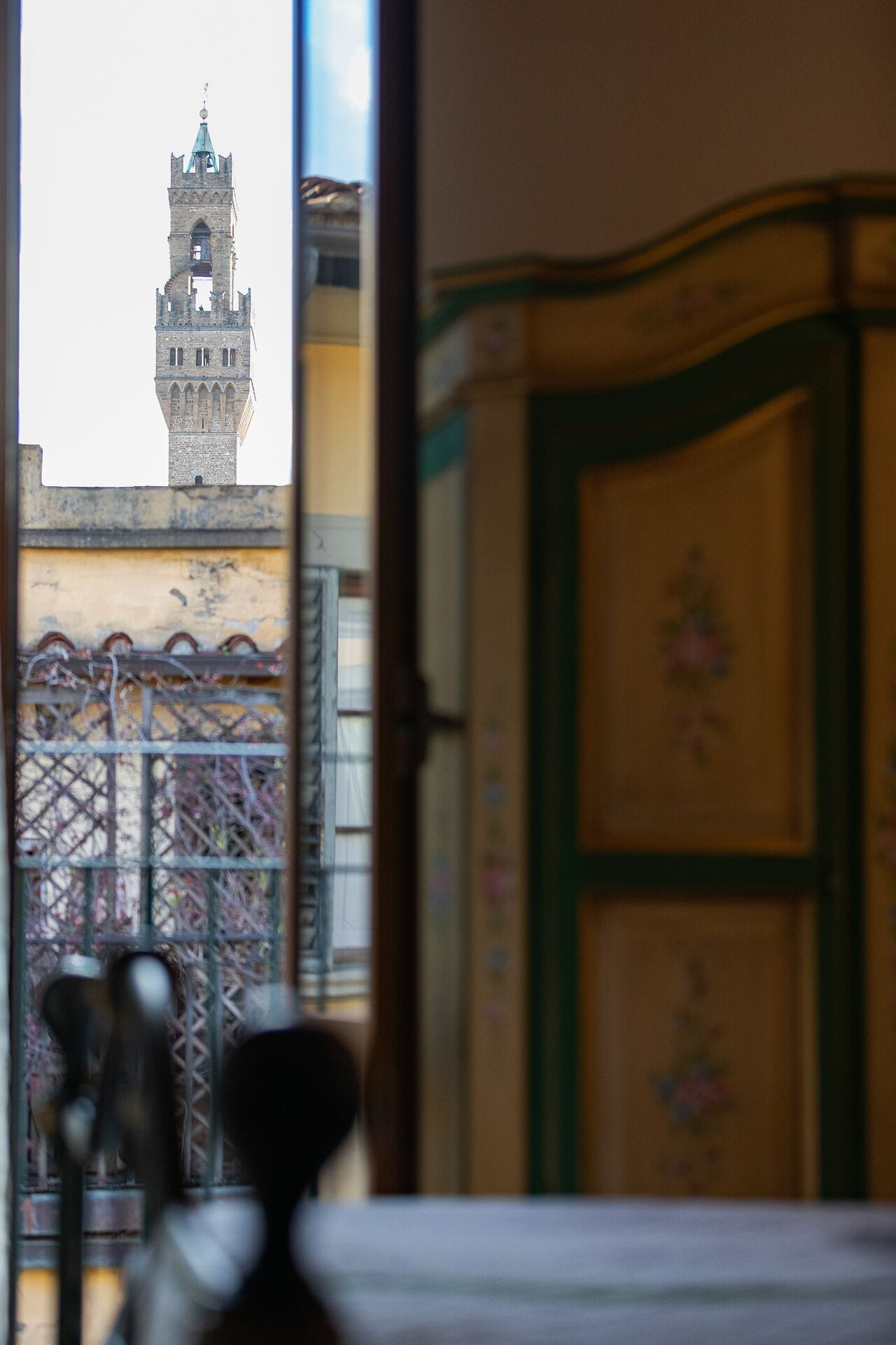 A view of a historic tower through a window, with a balcony and a decorative cabinet in the foreground.