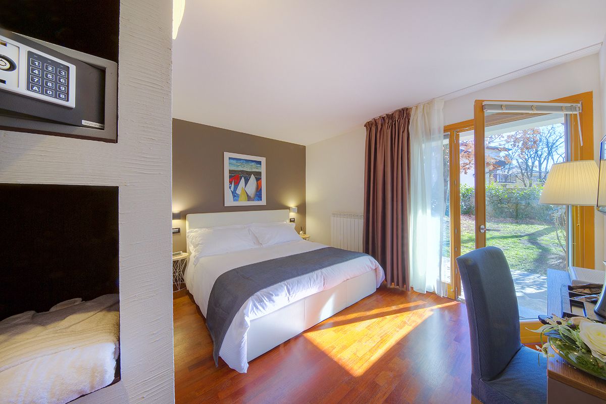 Habitación de hotel con cama, escritorio con lámpara, silla, ventana con cortinas y un cuadro en la pared.
