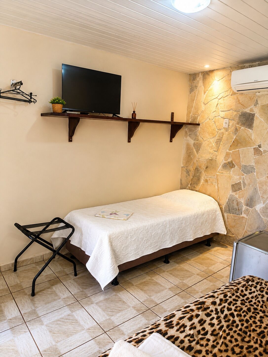A small room with a bed, a television mounted on a shelf, a stone wall, and a tiled floor.