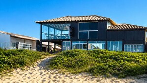 Ein Anwesen direkt am Strand mit überdachter Terrasse, einem großen, fensterdurchfluteten Gebäude und einem Sandweg, der zum Eingang führt.