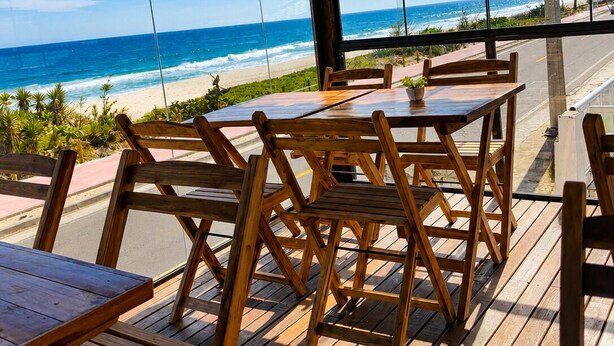 Ein Sitzbereich im Freien mit Holztischen und -stühlen mit Blick auf Strand und Meer.