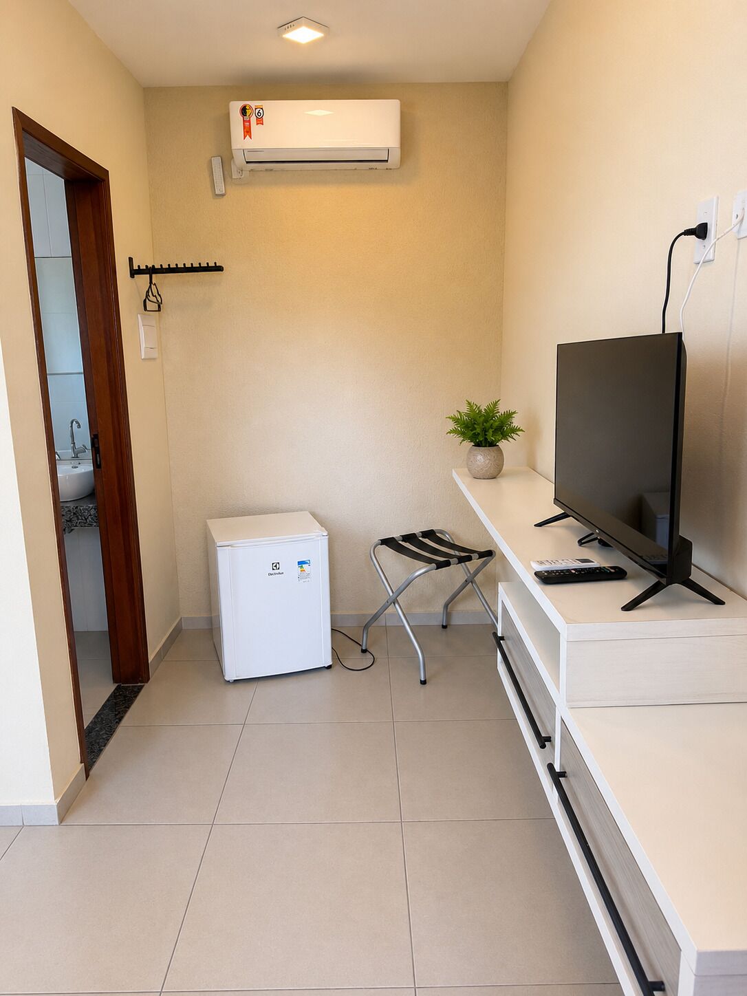 A compact room with a white air conditioner, a small refrigerator, a desk with a computer and keyboard, a chair, and a potted plant.