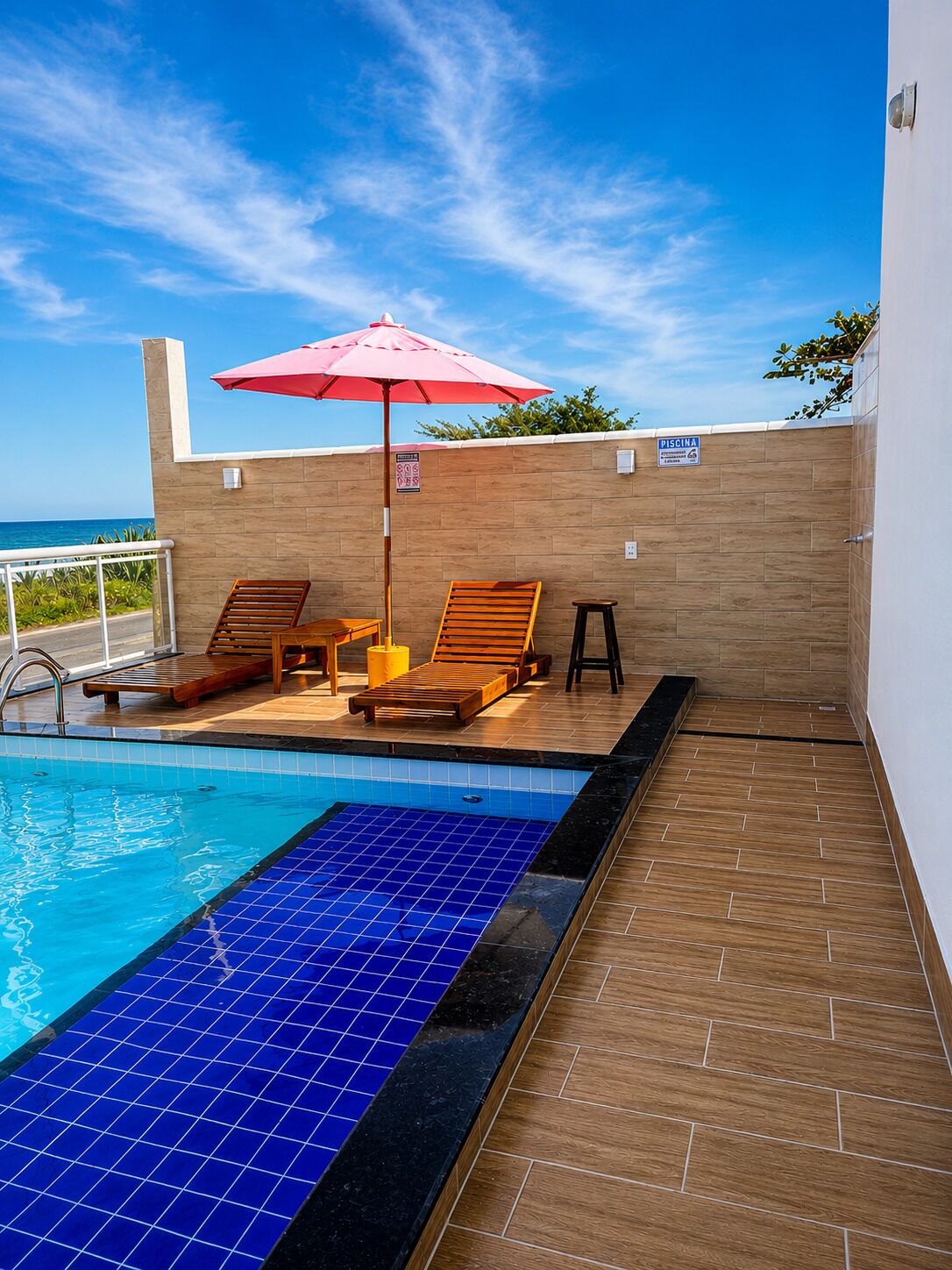A swimming pool with lounge chairs and an umbrella, a tiled deck, and a view of the ocean.