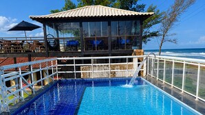 Ein Pool mit gefliestem Deck, eine Lounge mit Glasfassade und Blick auf den Ozean.