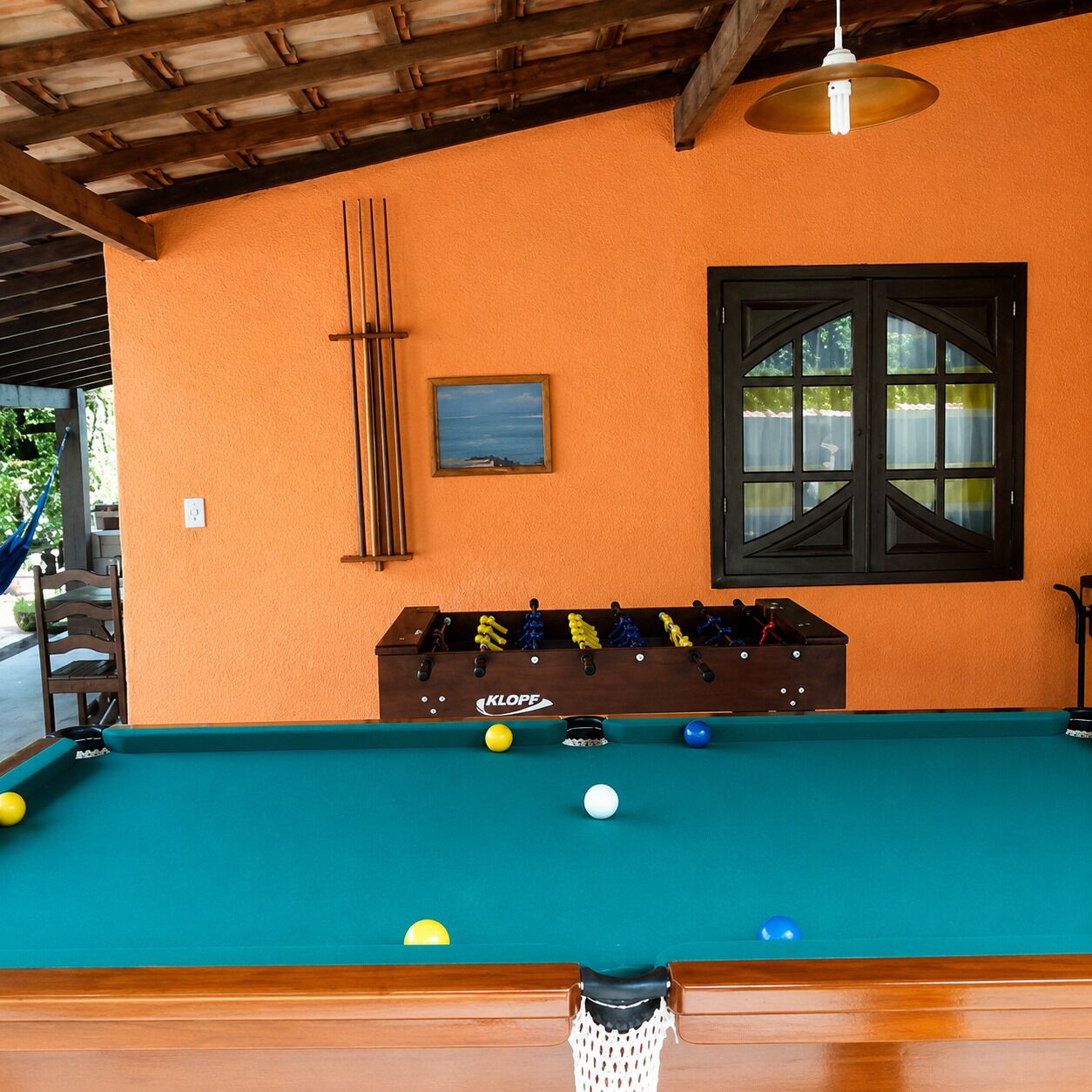 A pool table with balls, a foosball table, a window with curtains, and a hanging light fixture.
