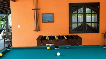 A pool table with balls, a foosball table, a window with curtains, and a hanging light fixture.