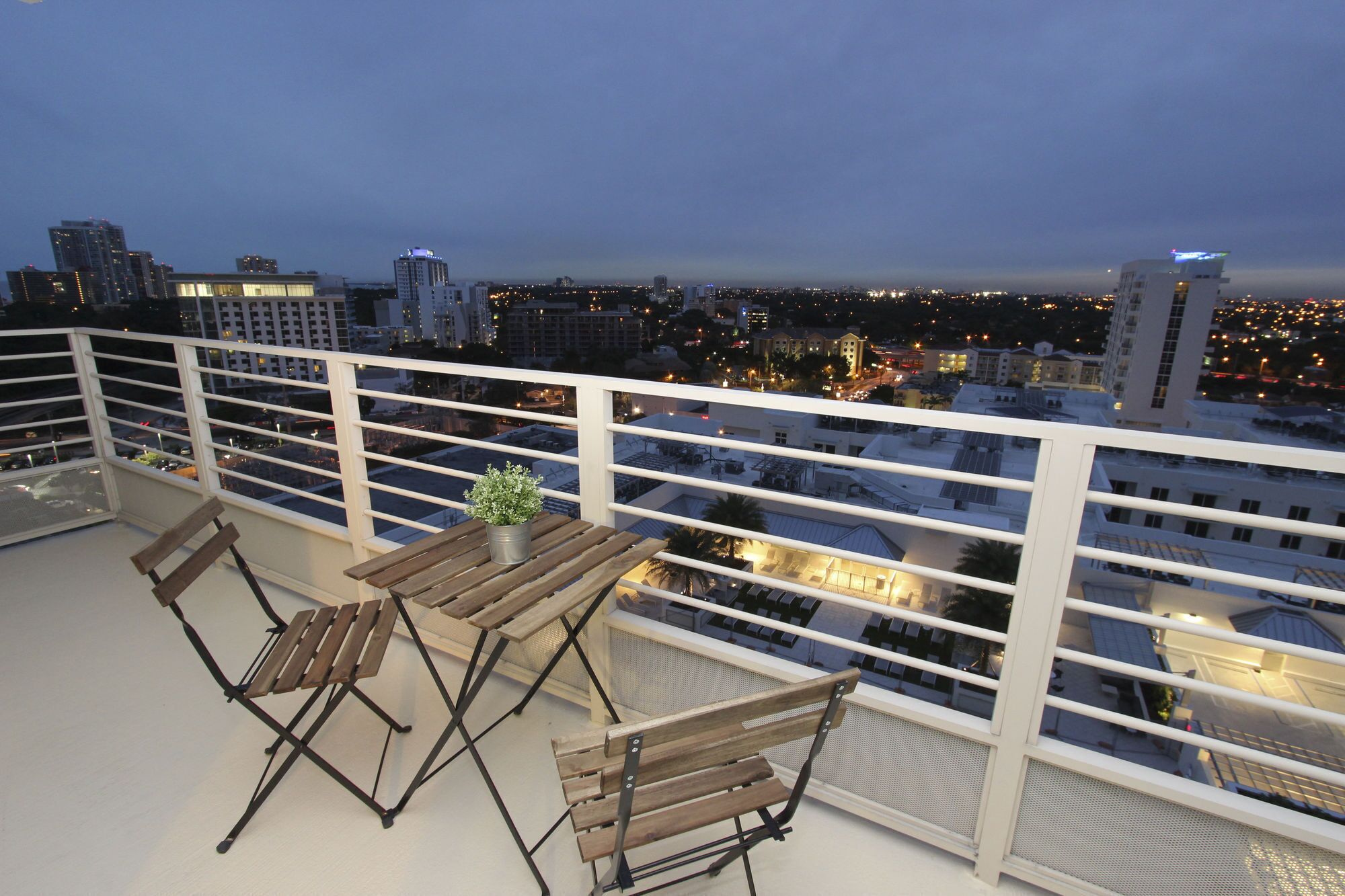 A balcony with a wooden table and chairs, overlooking a cityscape at dusk.