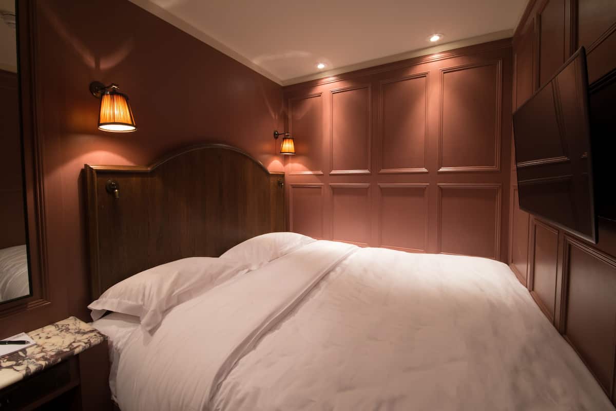 Un dormitorio con cabecera de madera, cama con ropa de cama blanca, mesita de noche con lámpara y televisor montado en la pared.