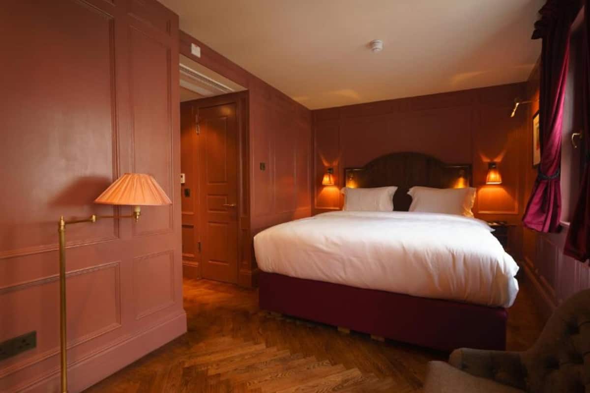 Una habitación de hotel con una cama grande, dos lámparas de noche, suelo de madera y una puerta que da a otra habitación.