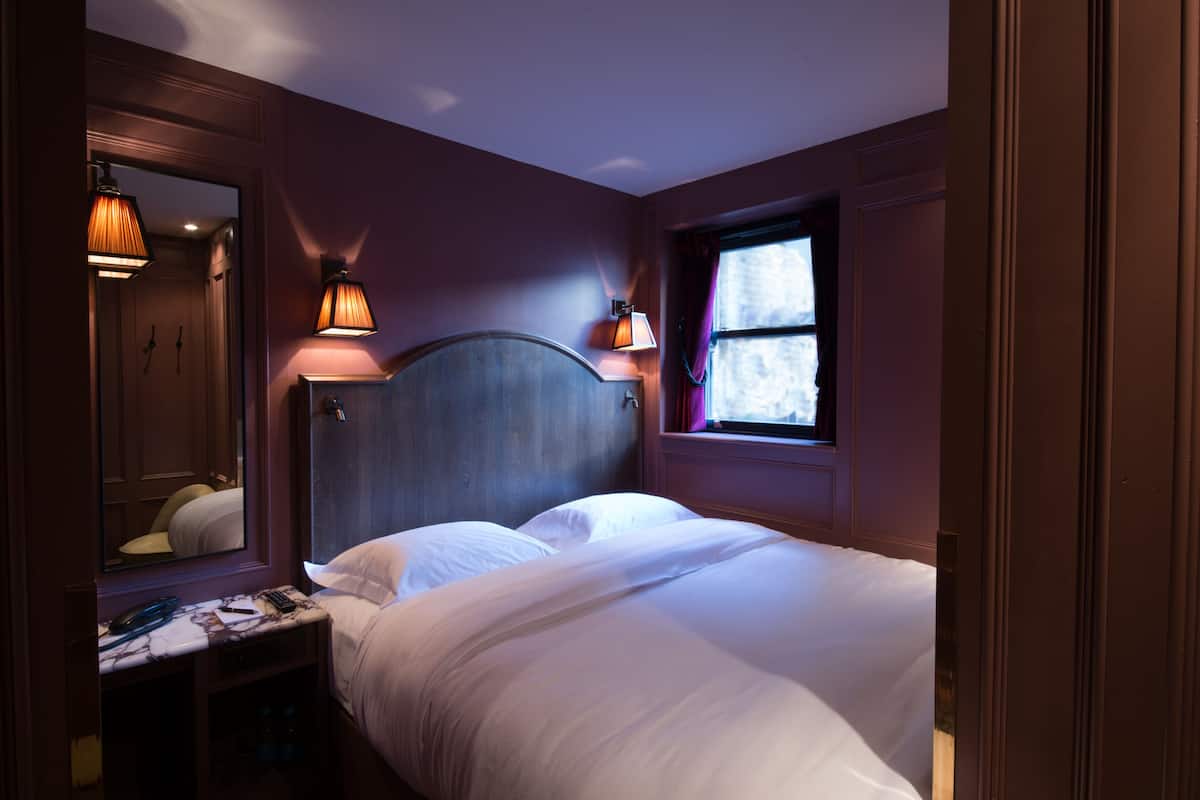 Habitación de hotel con una cama grande, una mesita de noche con lámpara, un espejo y una ventana con cortinas.