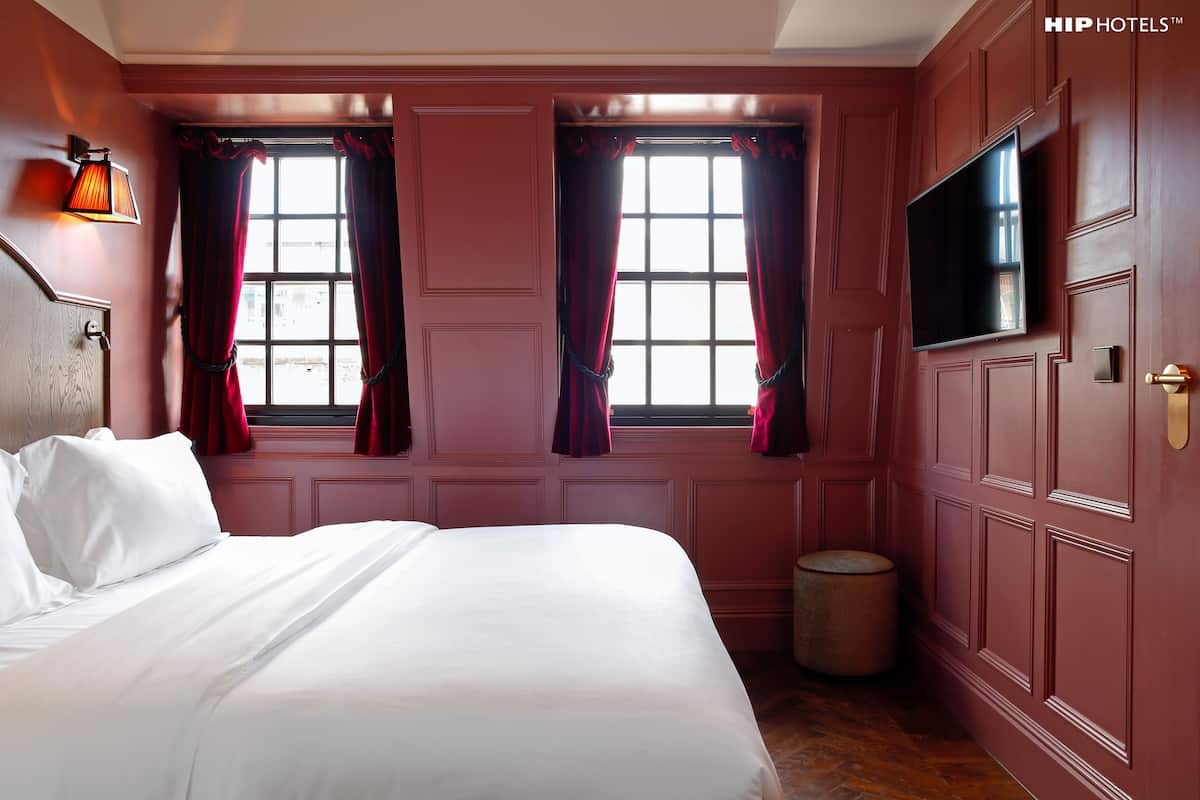 Habitación de hotel con una cama grande, dos ventanas con cortinas rojas, un televisor en la pared y suelo de madera.
